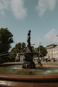 View of the Havis Amanda fountain statue with water flowing into the basin, surrounded by trees and historic buildings in Helsinki, Uusimaa, Finland.