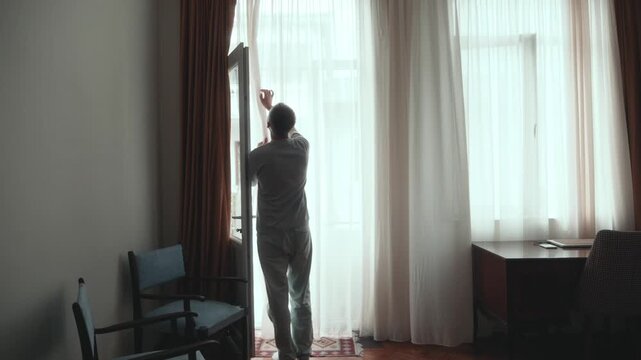 Man looking out window, pulling curtain aside and peering toward street from balcony doorway, checking outside at home in quiet solitary moment, slow motion.