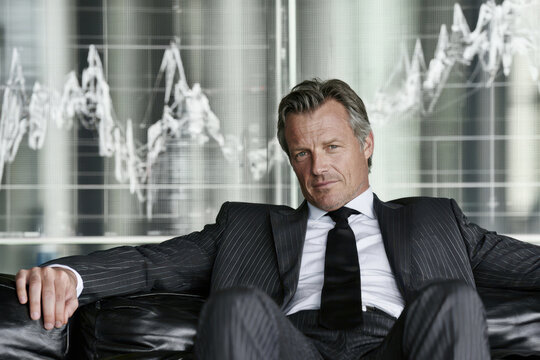Mature businessman in pinstripe suit seated on leather sofa with arms outstretched before financial charts on glass wall in modern office interior