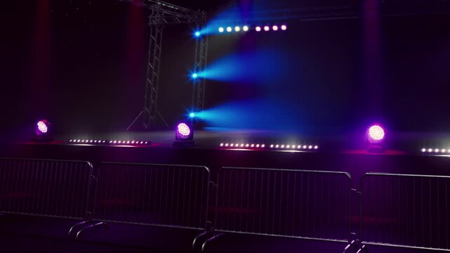 Colorful lights illuminate an empty stage, creating an electric atmosphere. Barriers stand waiting for an eager crowd as anticipation builds for the upcoming show.