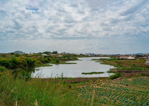 View of a tranquil lake surrounded by lush green vegetation, agricultural fields, and distant hills under a cloudy sky in Jos South, Plateau, Nigeria.