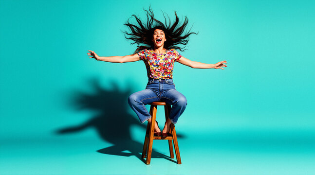 Joyful woman in colorful top and jeans on stool, turquoise background, pop-art. Dynamic studio lifestyle image for fashion, sales, energetic ads, and playful branding.
