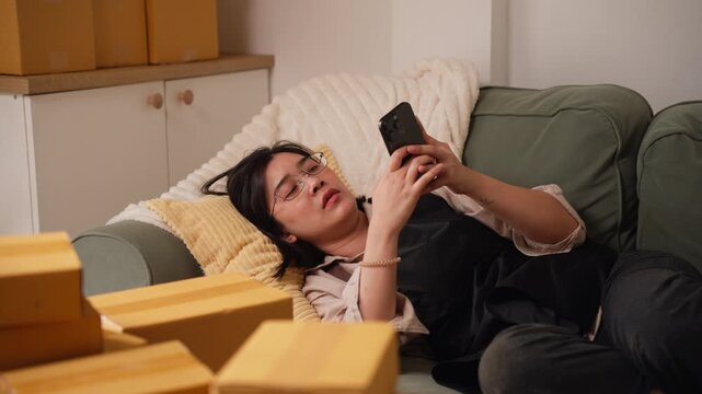 Exhausted Asian female merchant lying on couch holding smartphone with stressed face, suffering from burnout and heavy workload of sales.