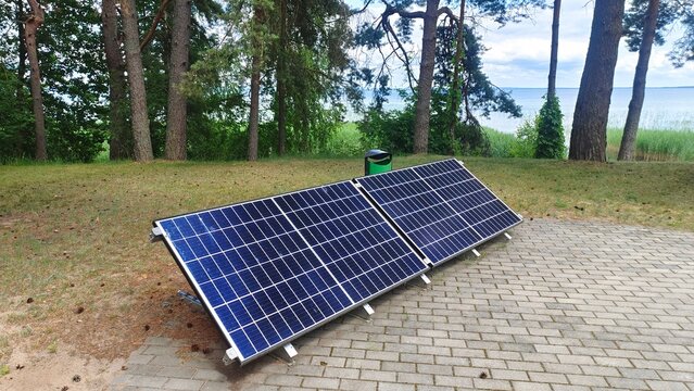 Solar panels for generating electricity are installed on on the tiled area. Nearby, on a grassy clearing strewn with pine cones and needles, grow pine trees, and beyond them lies a lake. Reeds