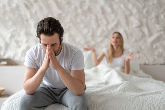 Worried Expression On Face. White Male In His 30s Sitting On Bed With Hands On Face, Listening To Arguments From Female Partner In Bedroom. Conflict And Relationship Issues Concept.
