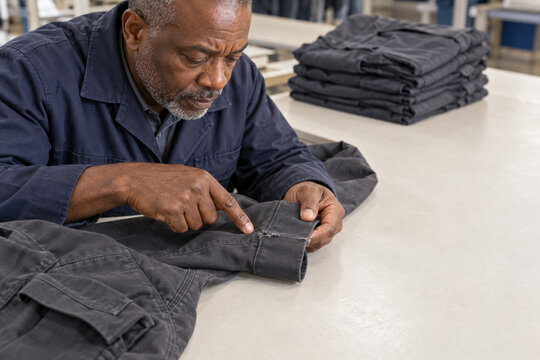 Adult man examines stitching on dark workwear trousers at a garment production table, with folded clothing in the background. Quality control concept.