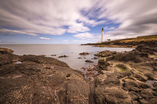 View of Scurdie Ness Lighthouse standing on a rocky coastline with long exposure clouds in the sky and calm sea in Montrose, Scotland, United Kingdom.