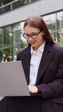Young businesswoman sitting on a bench in a city park, typing on a laptop and working outdoors while focused on the screen. Slow motion