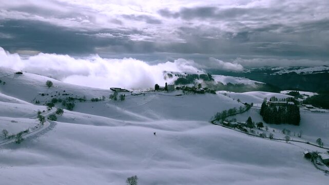 Aerial drone approach to small snowy mountain village emerging from clouds and fog, winter landscape with hills and dramatic sky