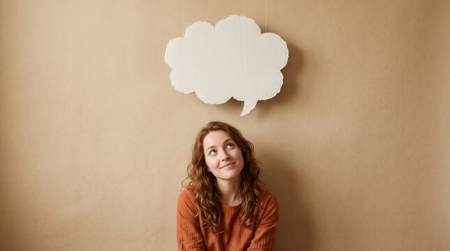 Young woman with curly hair looking up at a blank white thought bubble hanging against a plain beige background
