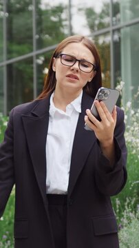 Young Caucasian woman walking in a city park, looking disappointed and concerned as she reads an unexpected message on her smartphone. Slow motion