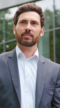 Handsome bearded businessman sitting on a bench in a city park, working on a laptop outdoors, focused on the screen before briefly looking away to the side. Slow motion