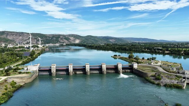 Hydroelectric barrage standing across a wide river, regulating water flow through gates amidst green shores and distant hills under a blue sky on a sunny summer day
