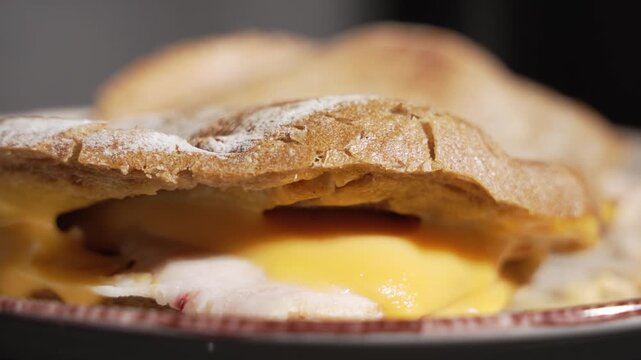 Low angle close-up of a toasted sandwich with oozing melted yellow cheese and sliced meat on a plate against a dark background, with slow camera movement.