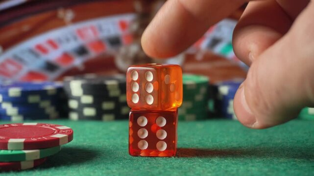 Hand placing translucent orange dice on a stacked pair beside poker chips on green casino table, with blurred playing cards and chips in the background
