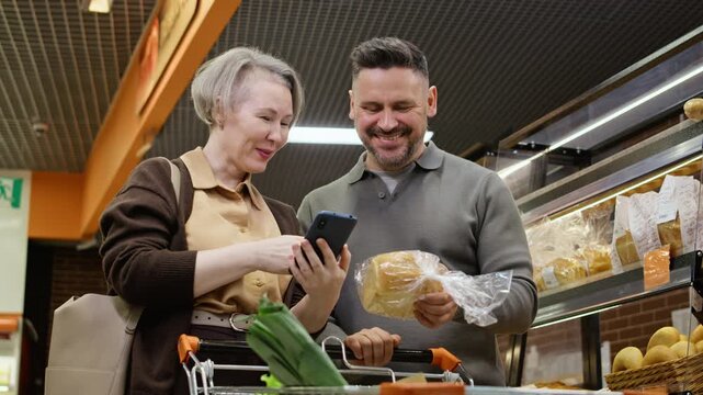 Middle aged Caucasian couple using smartphone while buying bread in supermarket bakery. Ideal for grocery delivery applications, retail marketing, and consumer lifestyle campaigns