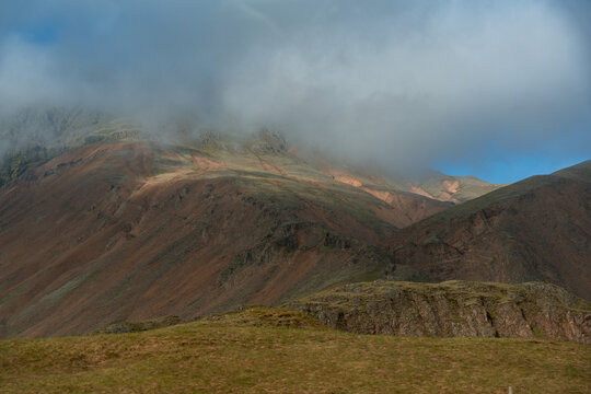 View of rugged volcanic mountains partially covered by low-hanging clouds and mist with grassy slopes in the foreground, Iceland.