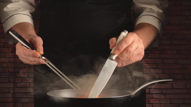 A cook holds pieces of pumpkin with a fork and guides them with a knife into a pan from which steam rises, demonstrating the cooking process against the backdrop of a dark brick wall