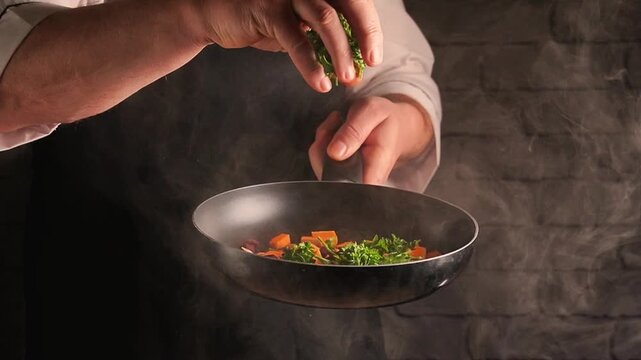 Cook adding fresh chopped herbs to steaming saute pan with diced vegetables in a dark kitchen setting
