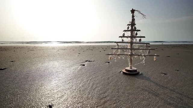 Small wooden christmas tree decorated with seashells standing on a wet sandy beach as gentle ocean waves wash ashore during a beautiful sunny morning, representing a sustainable holiday