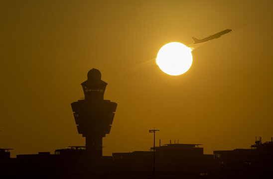 View of the Amsterdam Airport Schiphol control tower silhouetted against a vibrant orange sunset with an airplane taking off in the background in Schiphol, North Holland, Netherlands.