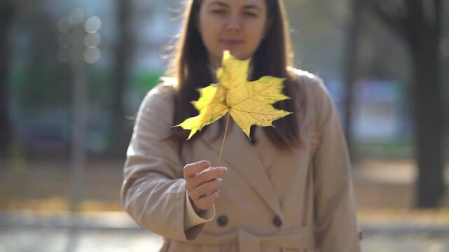 Woman holds a bright yellow maple leaf in autumn sunlight, soft blurred park background, calm outdoor moment.