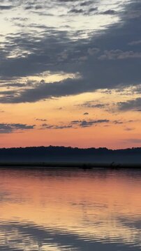 Time-lapse of gray clouds drifting over an orange sunset band on a river, vertical HD