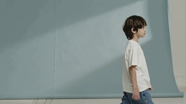 On cue walking child, white tee, blue jeans, crossing pale-blue seamed backdrop into angled light