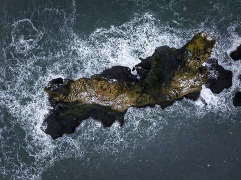 View of a mossy sea stack surrounded by crashing waves in the Atlantic Ocean, Iceland.