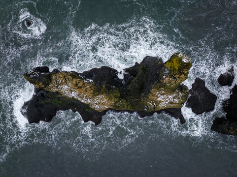 View of a rocky sea stack surrounded by crashing waves and white sea foam, Iceland.