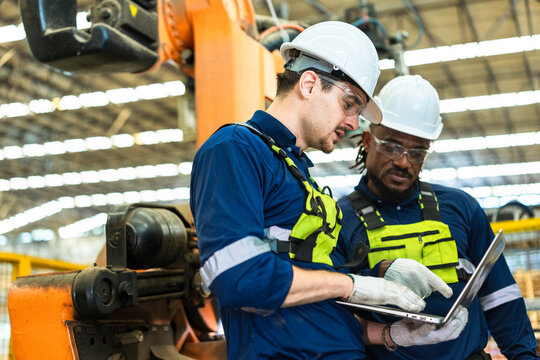 Two robotics engineers in white hard hats review a laptop by the robot arm as one points in a wood processing plant, a real wood manufacturing workplace in the AI era