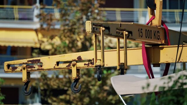 Rusty yellow crane lifting beam rated 15 000 kg with hooks and red straps hoisting a white boat hull in front of apartment buildings, French Riviera, France.