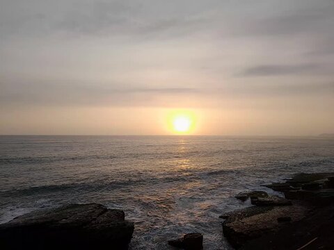 Warm evening sun orb descending over calm ocean waves along rocky coast horizon