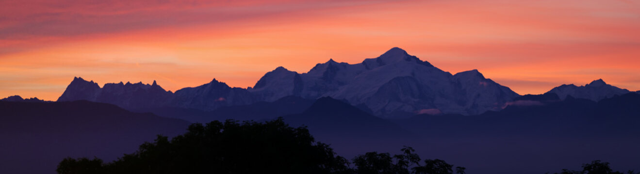 View of sunrise over the Mont Blanc massif with vibrant orange clouds above snow-capped peaks and dark tree silhouettes in Chamonix, Auvergne-Rhone-Alpes, France.