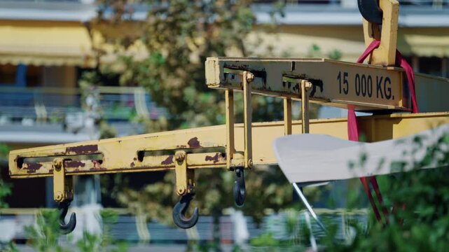 Yellow crane spreader beam with hooks and red lifting straps holding a white boat hull above green bushes and apartment balconies, French Riviera, France.