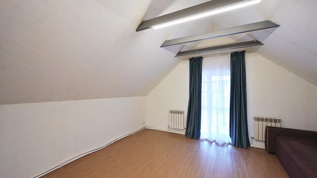 Spacious attic room with slanted ceiling, linear lights, window with dark and sheer curtains, wooden floor, partial brown sofa, radiators.
