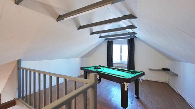 Attic billiard room with slanted ceiling, green table, wooden railing, exposed beams, window with dark curtains, minimalist finish.
