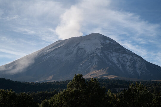 View of the snow-capped Popocatepetl volcano emitting smoke above a dense pine forest in Mexico.