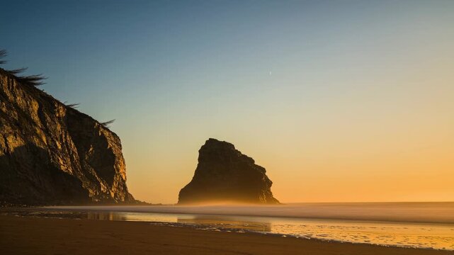 Standing lone sea stack catching shifting sunlight with sunlit cliff at left, mist and rolling surf