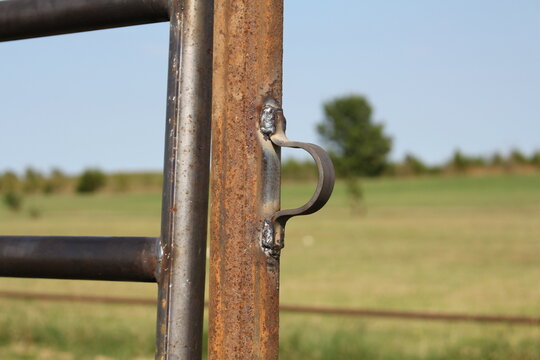 Welded hook on a rusty steel gate post with blurred pasture