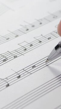 Hand writes musical notes on sheet music. Pen edits composition in close-up. Detailed notation practice in bright workspace supports music theory lessons, songwriting, and audio production.