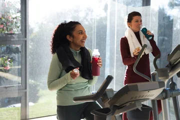 Diverse female friends using adjacent cardio machines in gym with towels, holding water bottles © WavebreakMediaMicro