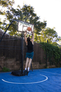 Male jumping near portable hoop, holding basketball, wearing black tank, teal shorts on blue court