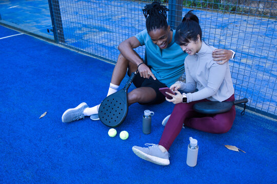 Diverse couple sitting on blue padel court near metal mesh fence, sharing smartphone with rackets