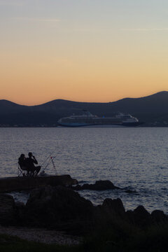 Two silhouetted anglers fish from a waterside pier beneath a glowing sunset sky over open water