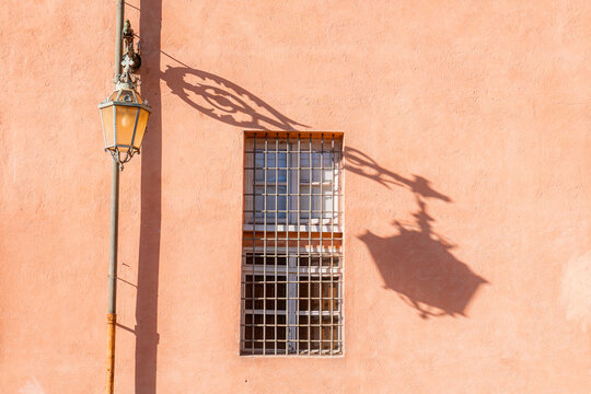 View of a vintage street lamp and its shadow cast on a peach-colored wall with a barred window in Turin, Piedmont, Italy.