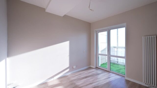 Spacious empty room with beige walls, wooden floor, large glass door, white radiator, minimalist interior, bright natural light.
