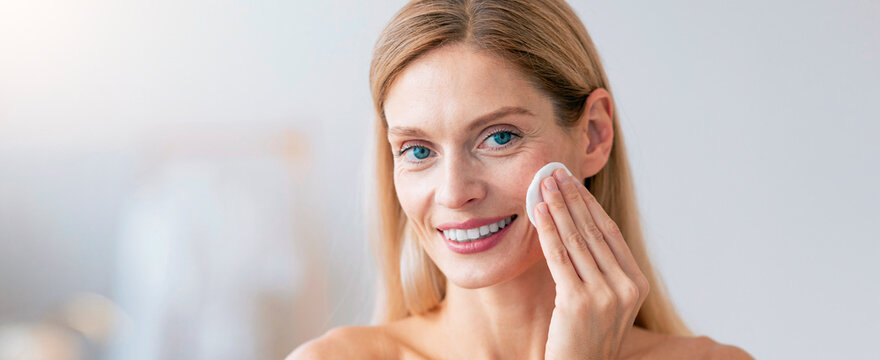 Happy Young Caucasian Woman Smiling While Applying Skincare Cream With Cotton Pad To Her Face, Standing In Bright Bathroom. Focused On Skincare Routine And Self-care, Home Setting.