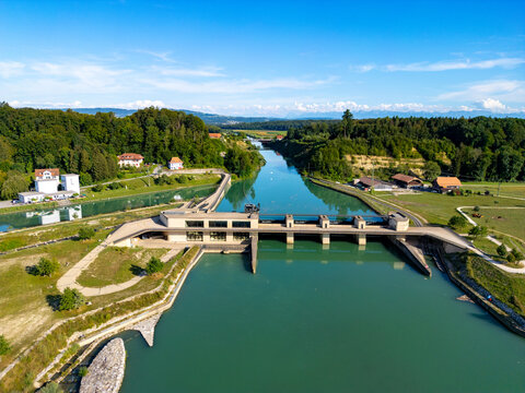 View of the Aare Seeland dam with concrete structures, calm turquoise river water, surrounding green fields, and dense forests, Aarberg, Canton of Bern, Switzerland.