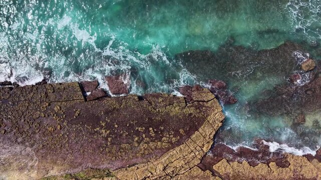 An aerial view of turquoise ocean waves crashing against a rocky shoreline